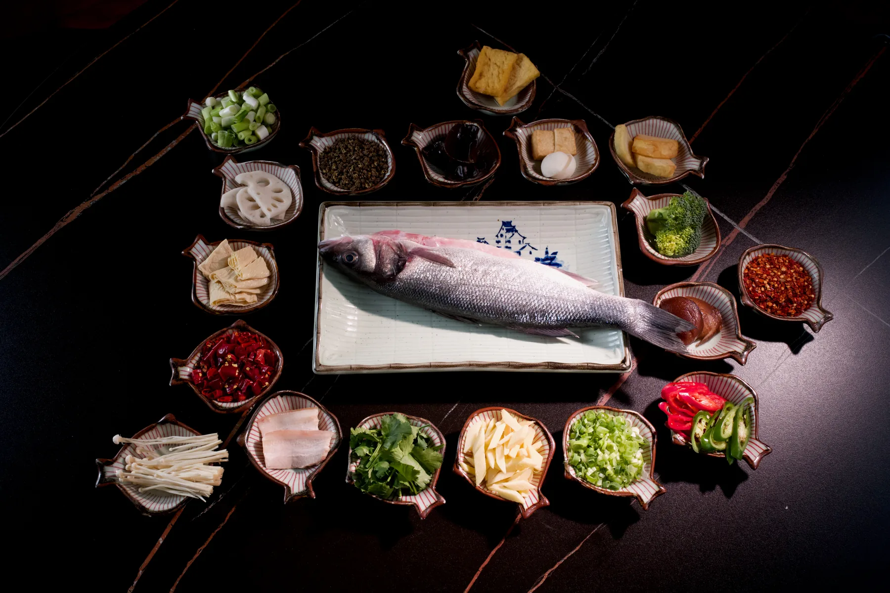 A whole raw sea bass on a long white plate, ringed by dishes of chilli, pickled greens, spring onion and bamboo