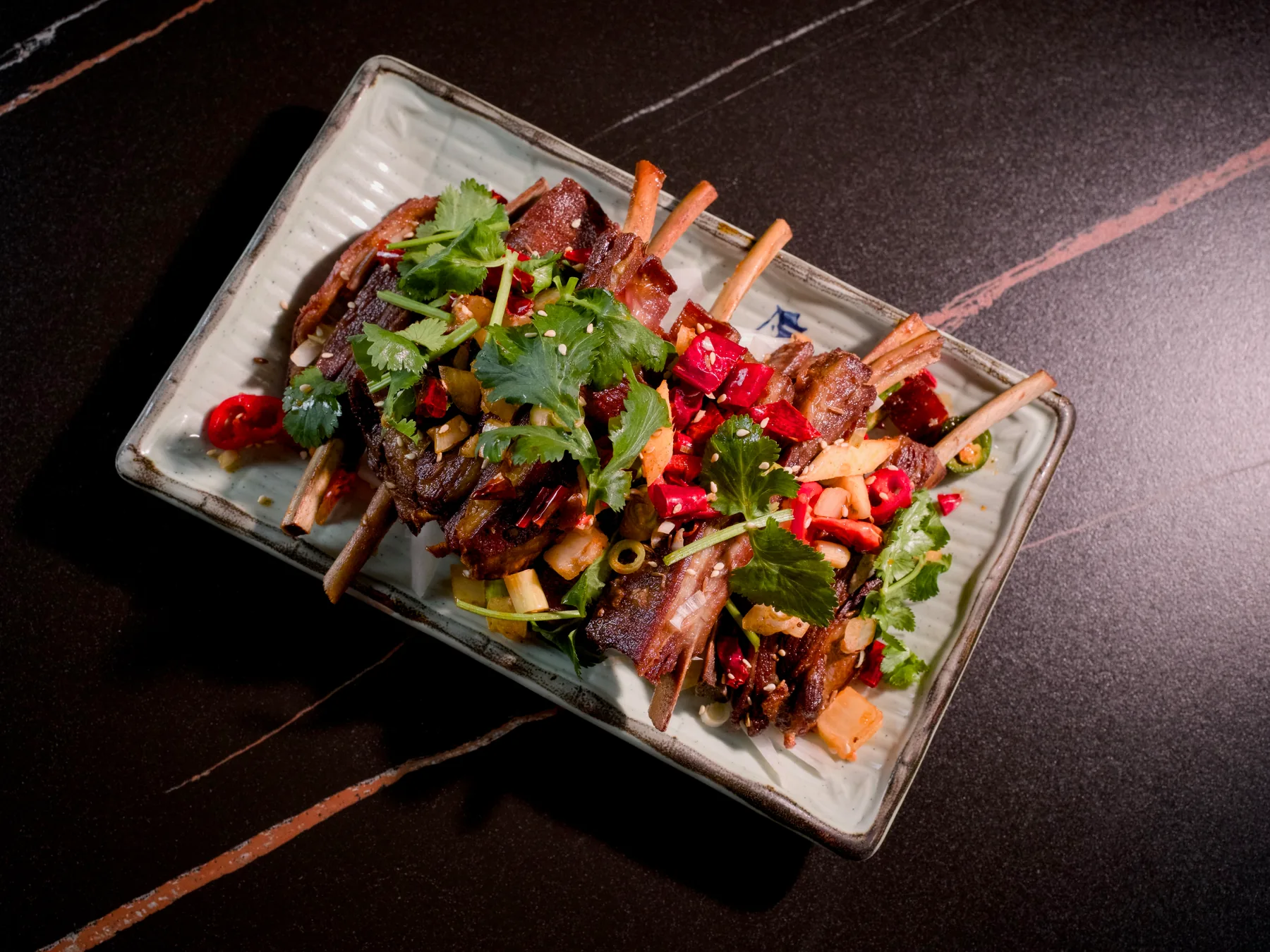 Grilled lamb chops on a long white plate under coriander, red chilli and toasted cumin