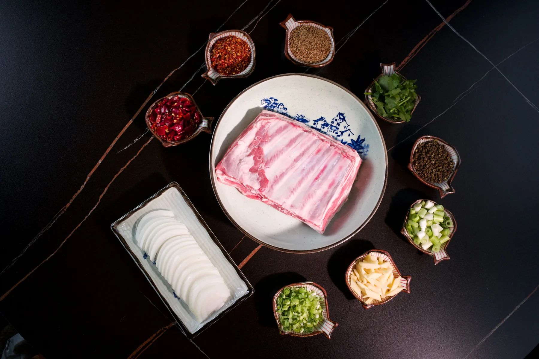 A raw rack of lamb on a white plate beside bowls of cumin, chilli flake, pomegranate and sliced garlic