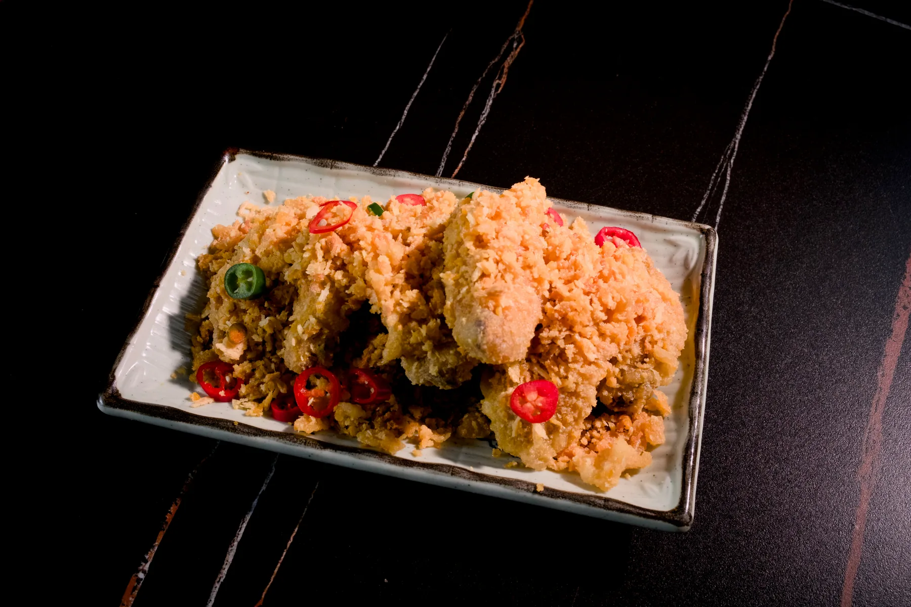 Chicken wings under a thick, sandy crust of fried salted egg yolk on a pale rectangular plate, scattered with sliced red and green chilli